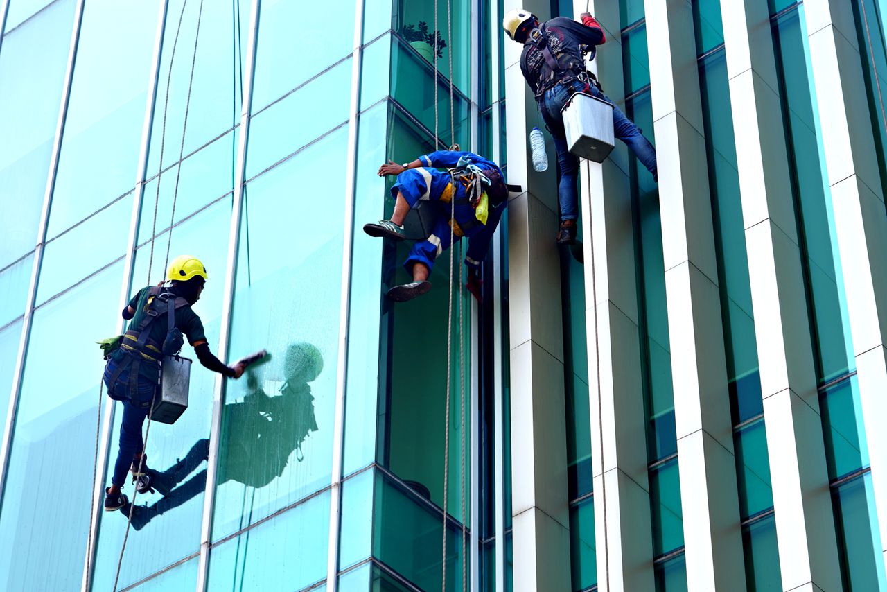 Trois nettoyeurs de vitres suspendus à des cordes nettoient, en journée, la façade d'un immeuble moderne en verre.